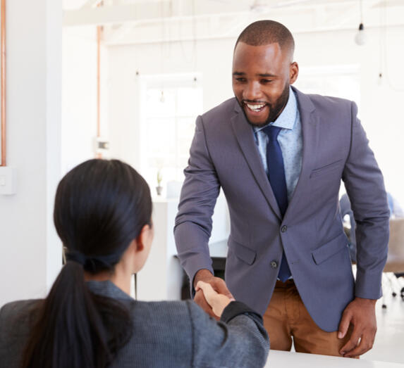 Image of a man in a suit standing while shaking the hand of a woman seated.